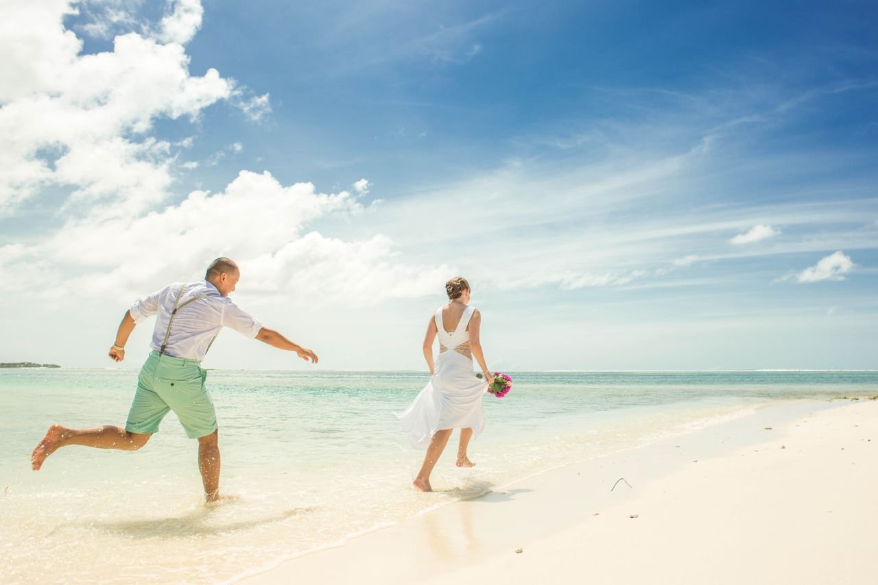 About A couple in wedding attire joyfully running along a sunny tropical beach.